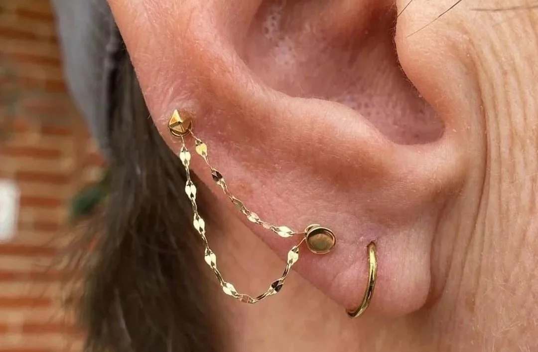 Close-up of a person's ear with three earrings: a gold hoop in the earlobe, a gold stud with a chain connecting to a second stud, and a small gold hoop in the helix.