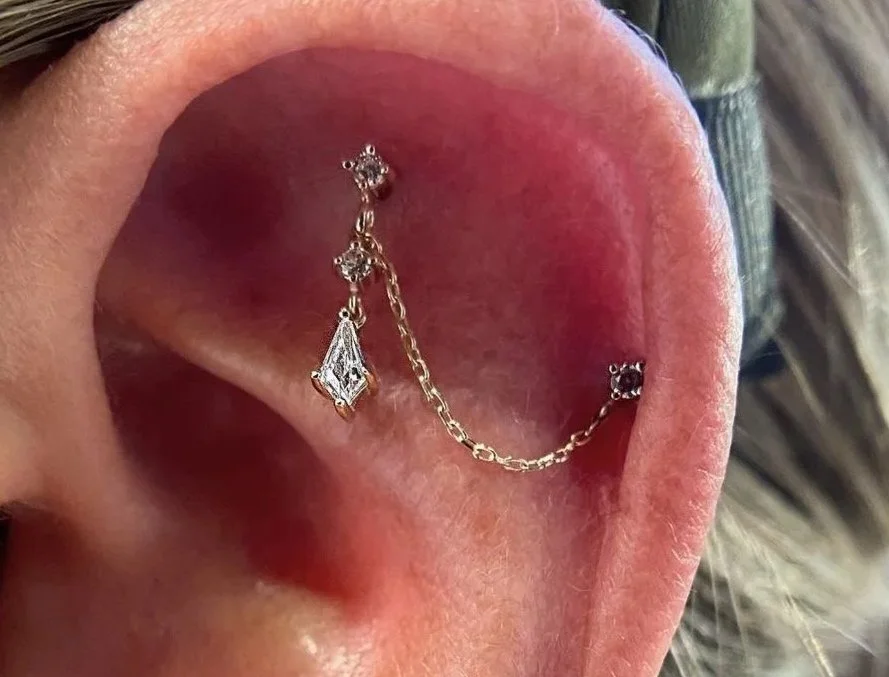 Close-up of a person's ear with multiple earrings, including a diamond stud, a decorative earring with a chain connecting two piercings, and a small gemstone stud.