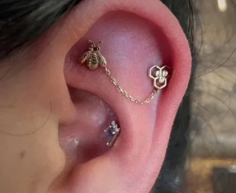 Close-up of a person's ear with multiple earrings, including a bee-shaped ear cuff connected to a flower-shaped stud, a small gemstone hoop, and a row of three small rhinestone earrings.