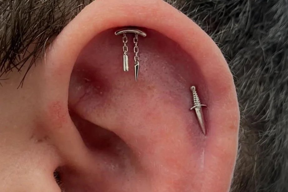 Close-up of a human ear with multiple piercings, including a chain with two charms and a dagger-shaped jewelry in the helix and a dagger-shaped earring in the lobe.
