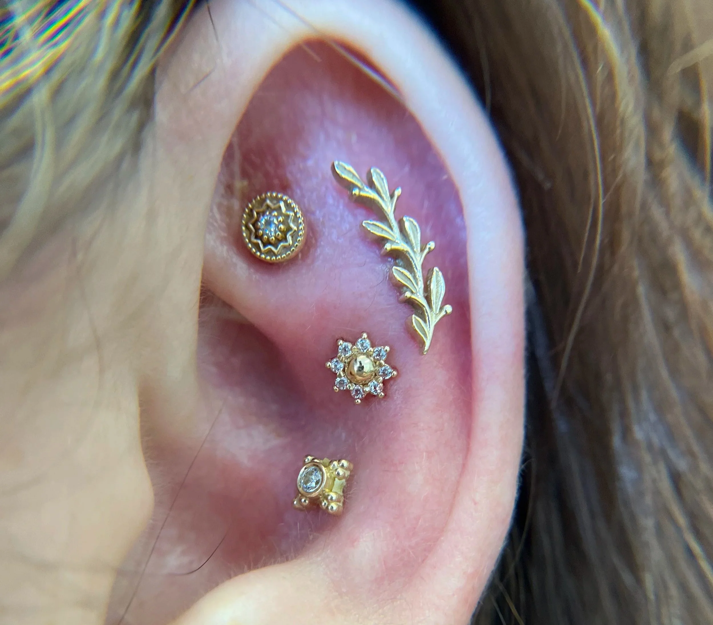 Close-up of a person's ear with multiple gold and diamond earrings, including a star-shaped stud, a leafy branch design, a curved bar with a small gemstone, and a square-shaped earring with a gemstone.