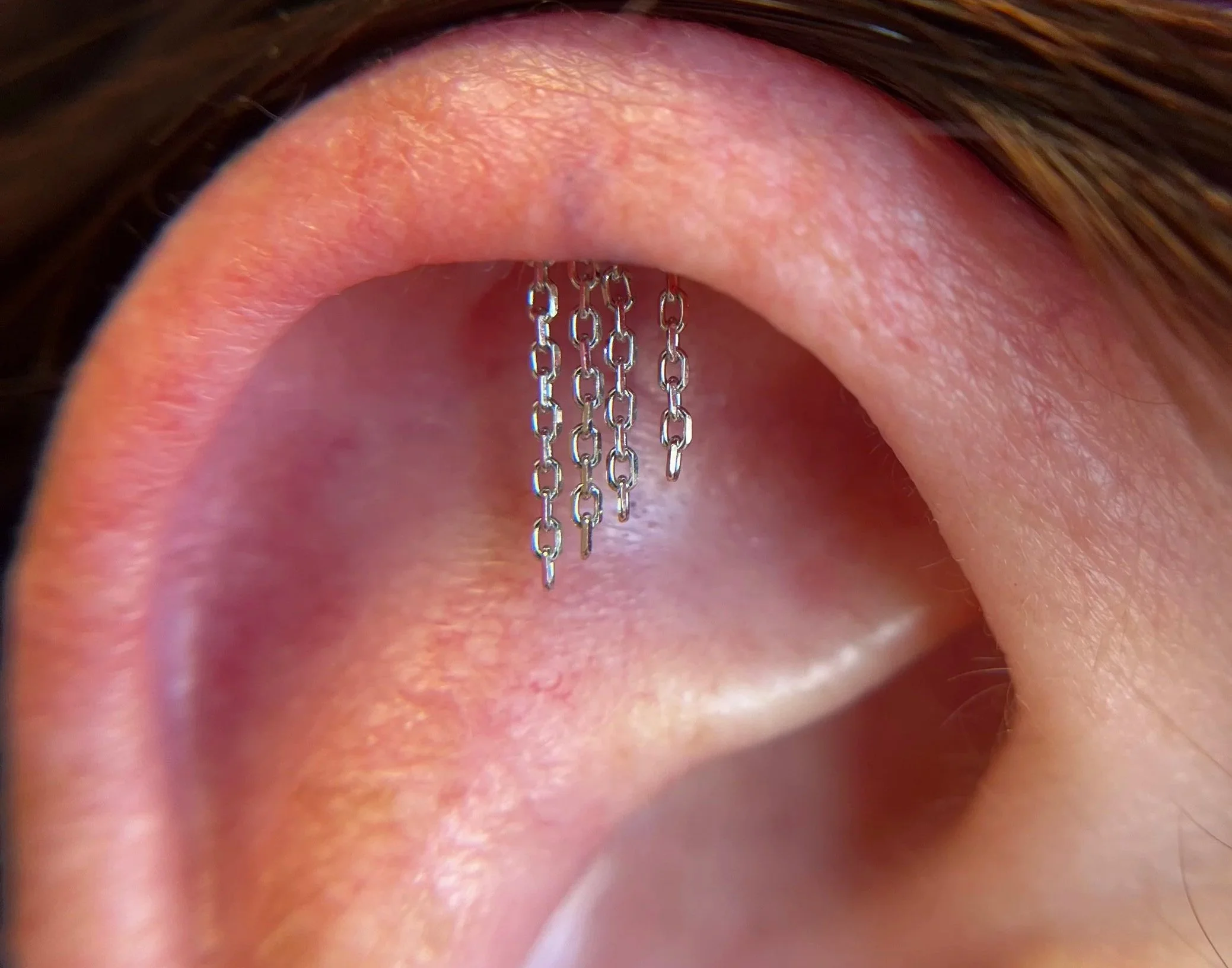 Close-up of a human ear with a small silver chain earring hanging from the upper ear, with brown hair partially covering the top of the ear.