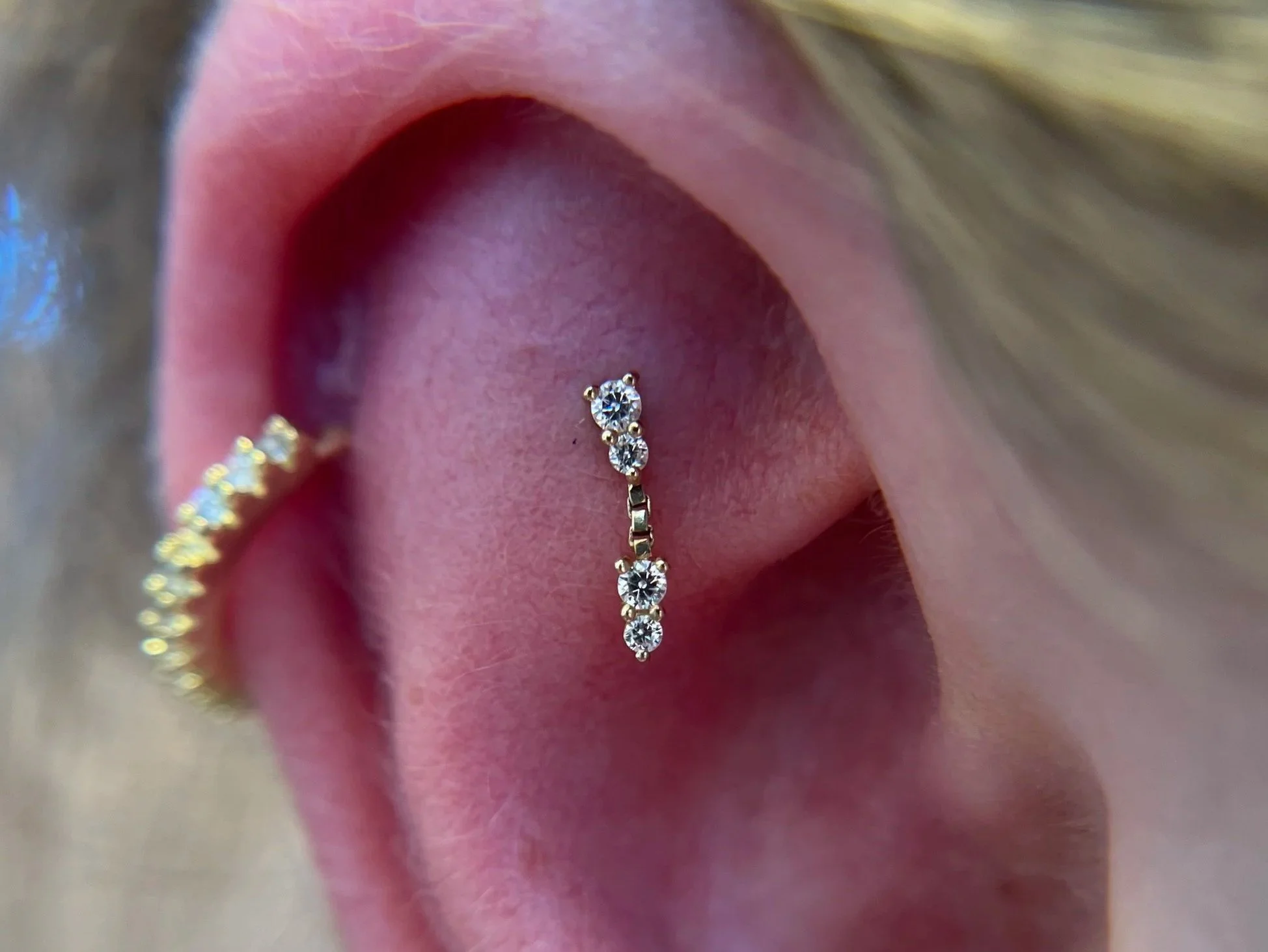 Close-up of a person's ear with multiple earrings, including a cartilage hoop with small diamonds, a vertically aligned row of diamonds in the earlobe, a shell-shaped earring, and a gold hoop with a blue gemstone.