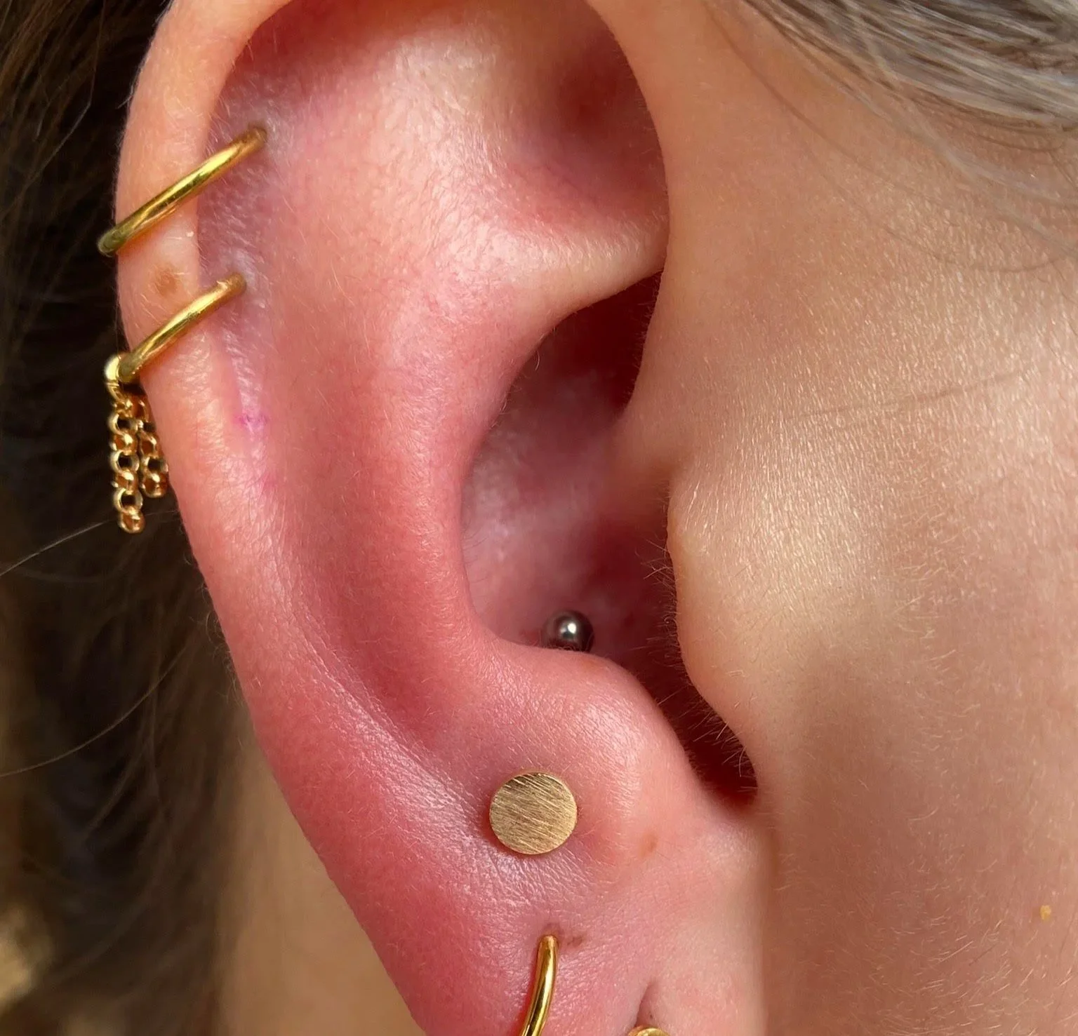 Close-up of a person’s ear with multiple gold earrings, including small hoops, a stud, and a cartilage earring with a dangling chain.
