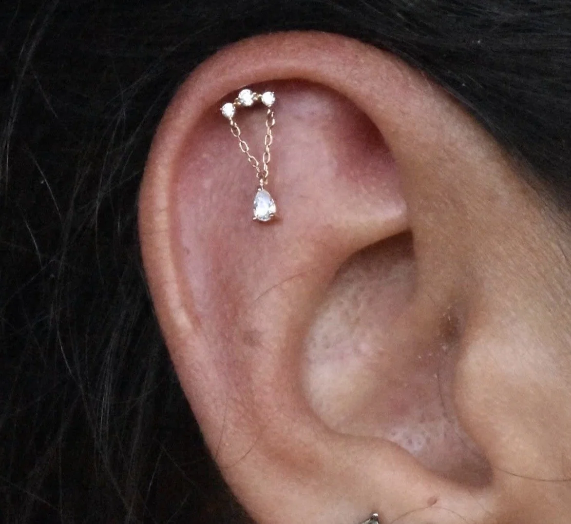 Close-up of a person's ear with a small, delicate gold chain and diamond drop earring piercing.