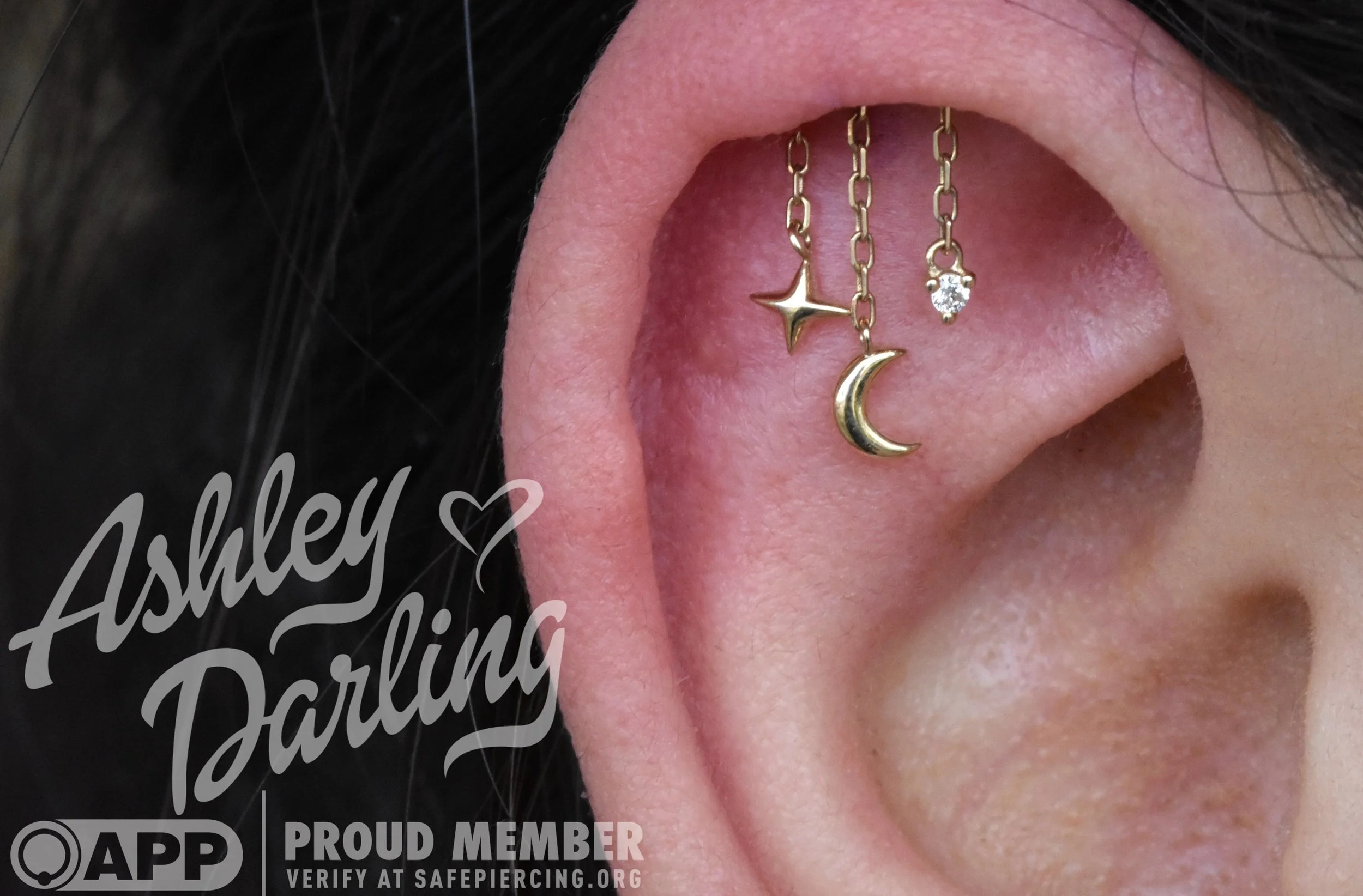 Close-up of a woman's ear with three gold earrings, including a star, moon, and diamond stud, and text overlay reading 'Ashley Darling' and 'Proud Member' from Safe Piercing.