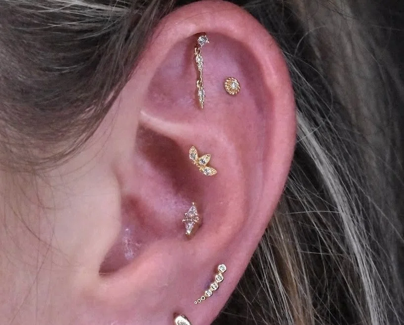 Close-up of a human ear with multiple gold and diamond earrings, including a long dangling earring, a small circular earring, a leaf-shaped earring, and a spiral helix and stud earrings.