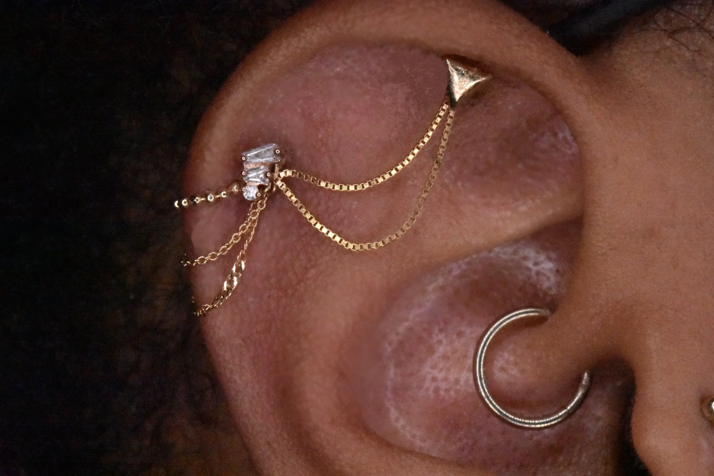 Close-up of a person’s ear adorned with multiple gold earrings and chains, with dark curly hair partially visible.