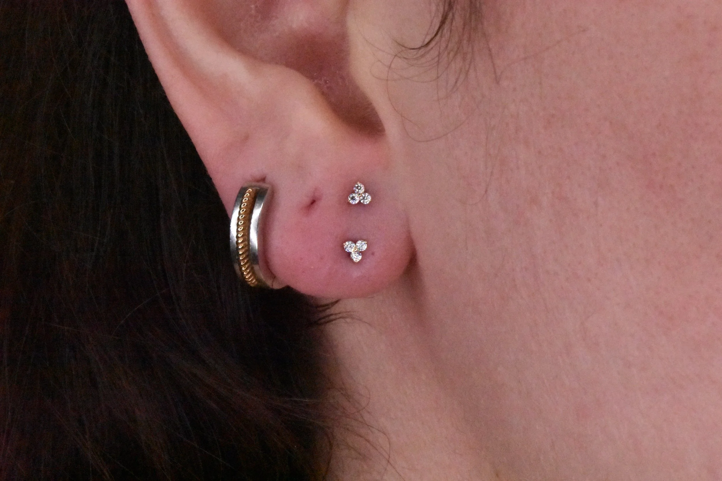 Close-up of a person's ear with multiple piercings, including small diamond flower-shaped earrings, a silver hoop, and a gold ring, with dark hair partially visible.