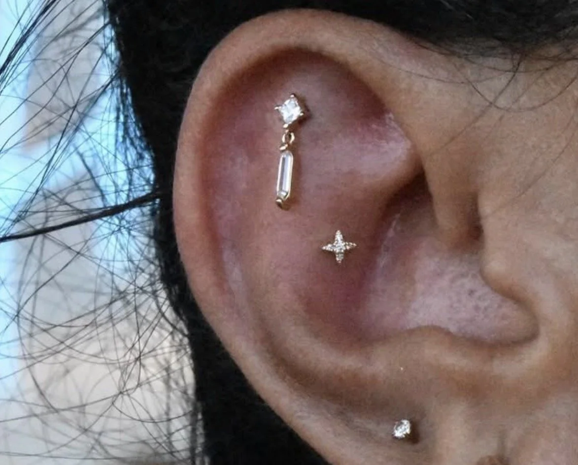 Close-up of a person's ear with multiple piercings, including a stud at the bottom, a star-shaped earring, and a gold barbell with a gemstone at the top.