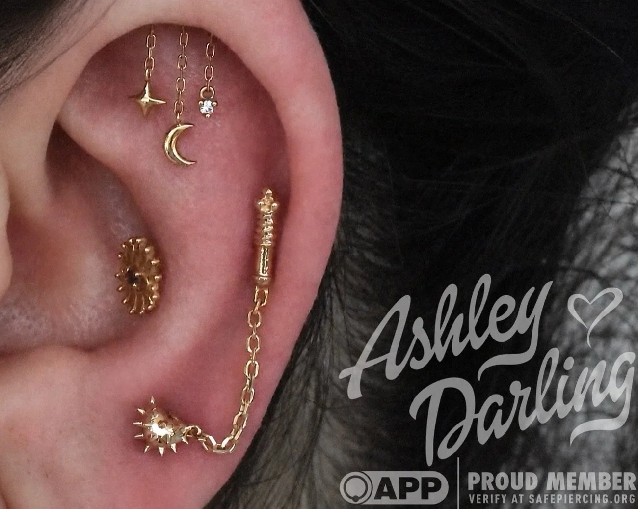 Close-up of a person's ear with multiple gold earrings including a moon, star, crystal, and spiked design earrings, with text reading 'Ashley Parking' and a heart, highlighting the jewelry.