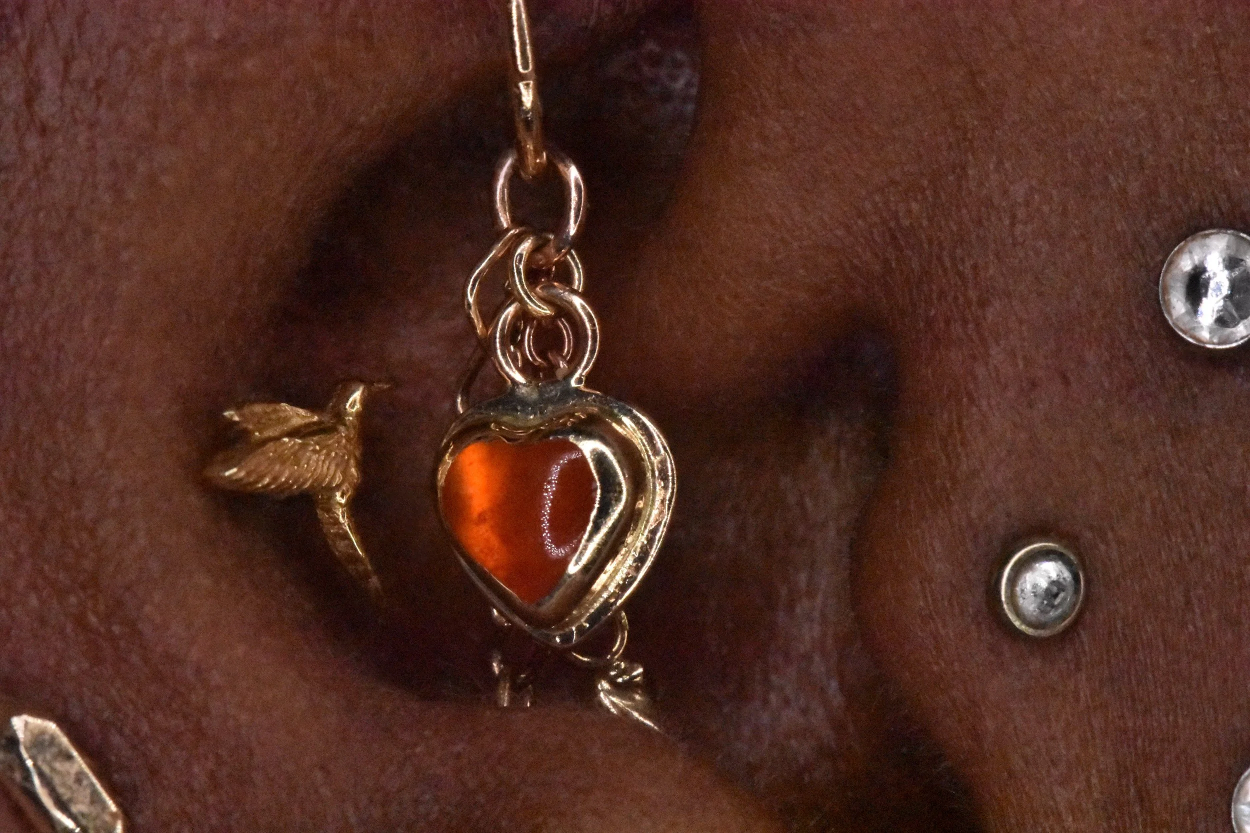 Close-up of a pierced earlobe with a heart-shaped jewelry piece containing an orange-red stone, accompanied by a gold-colored leaf charm hanging from the same jewelry, with clear rhinestones and a dark skin background.
