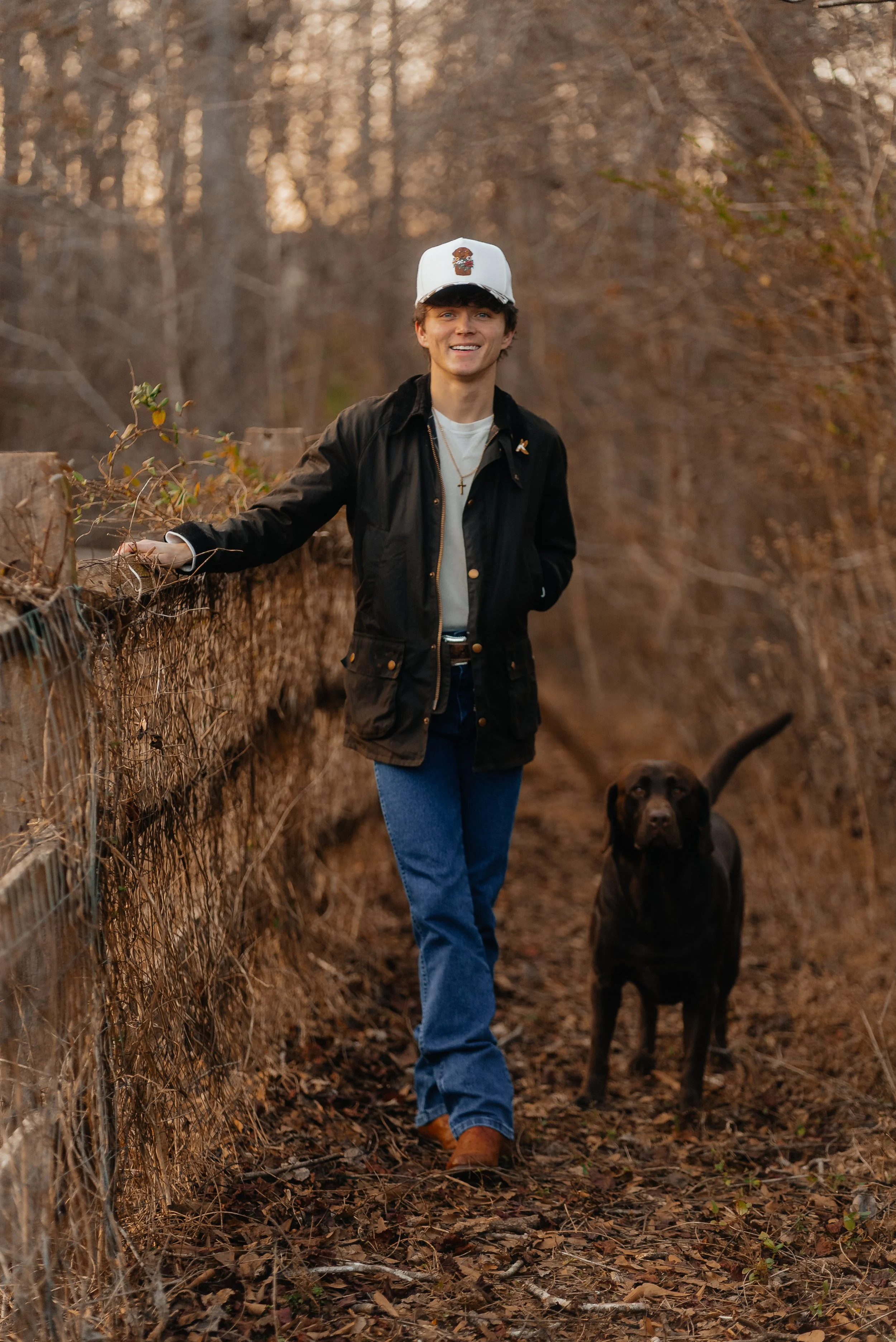 A young man with dark hair wearing a white cap, black jacket, white shirt, blue jeans, and brown boots, smiling and walking alongside a black Labrador retriever on a wooded trail during sunset.