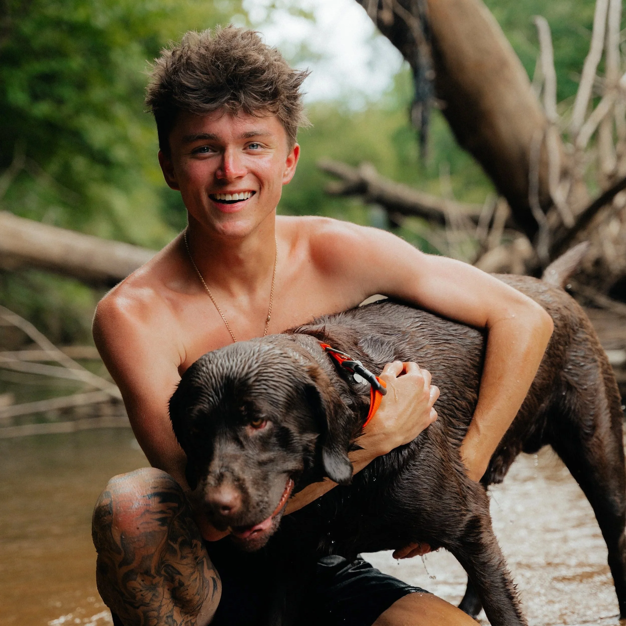 A young man with a big smile, short messy hair, and a tattoo on his knee, holds onto a wet, muddy dog with a red collar in a natural outdoor setting, likely near a body of water with trees in the background.