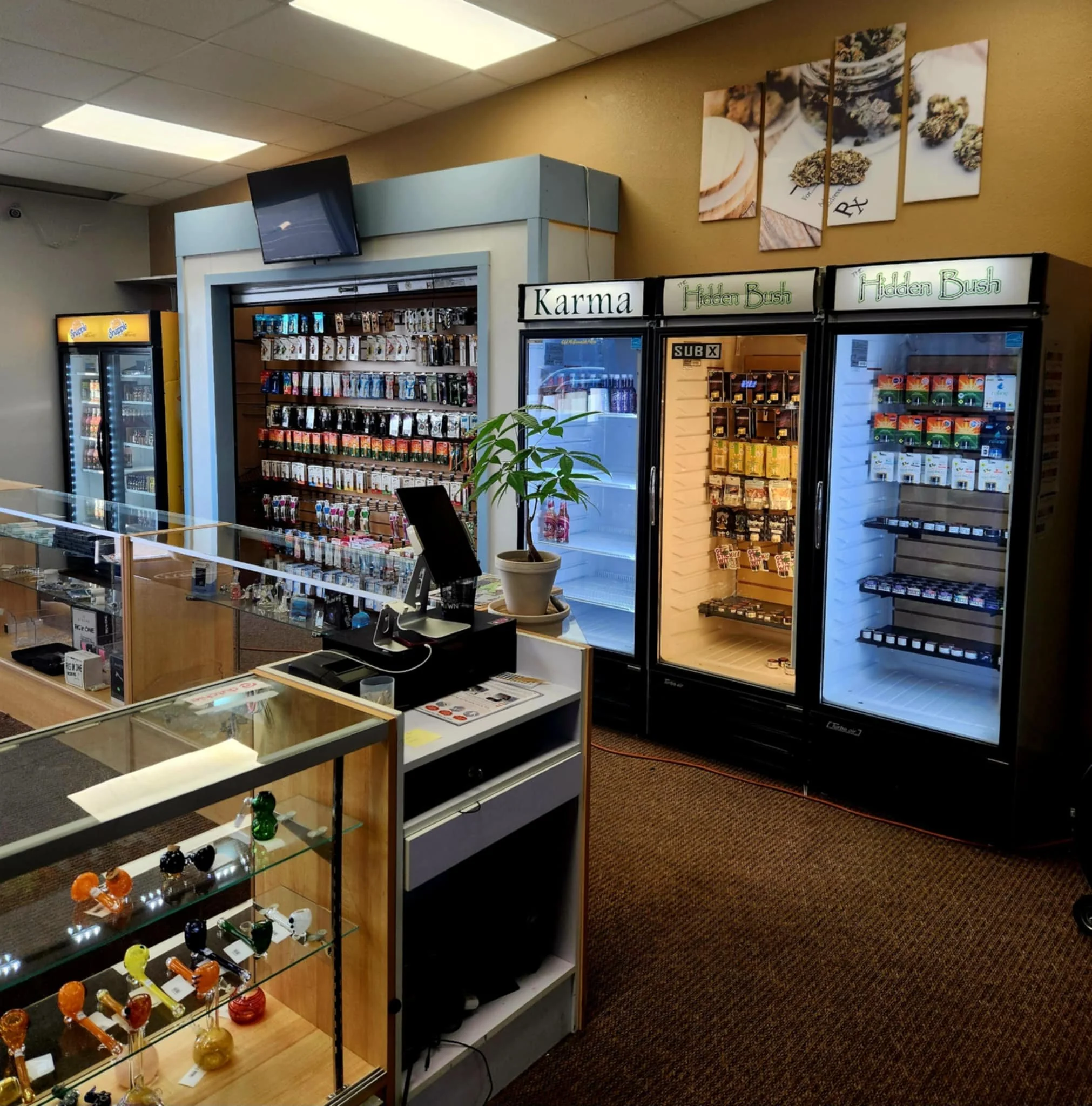 Interior of a retail shop with refrigerated display cases filled with various cannabis products, glass display cabinets containing colorful glass pipes, and a sales counter with a register.