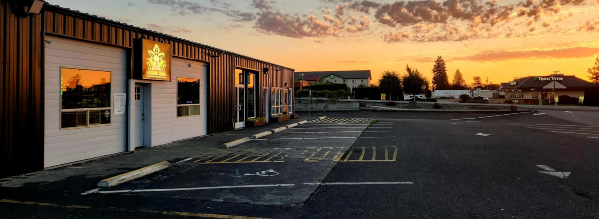 Commercial building and parking lot at sunset with orange sky and clouds, adjacent to another building with a Taco Time sign visible.