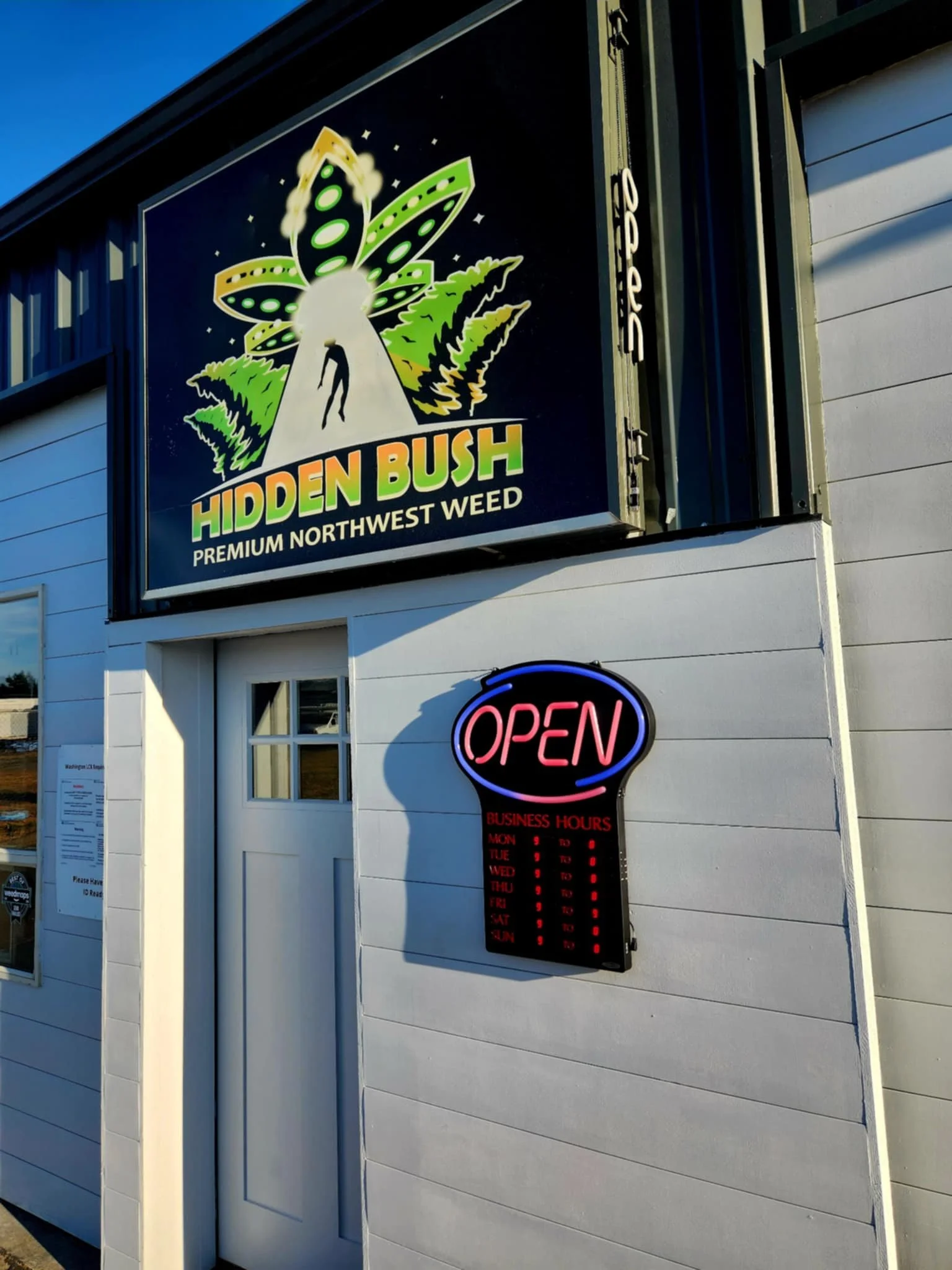 Storefront with a sign reading 'Hidden Bush Premium Northwest Weed,' featuring an alien-themed logo. Open sign with business hours displayed on the side of the building.