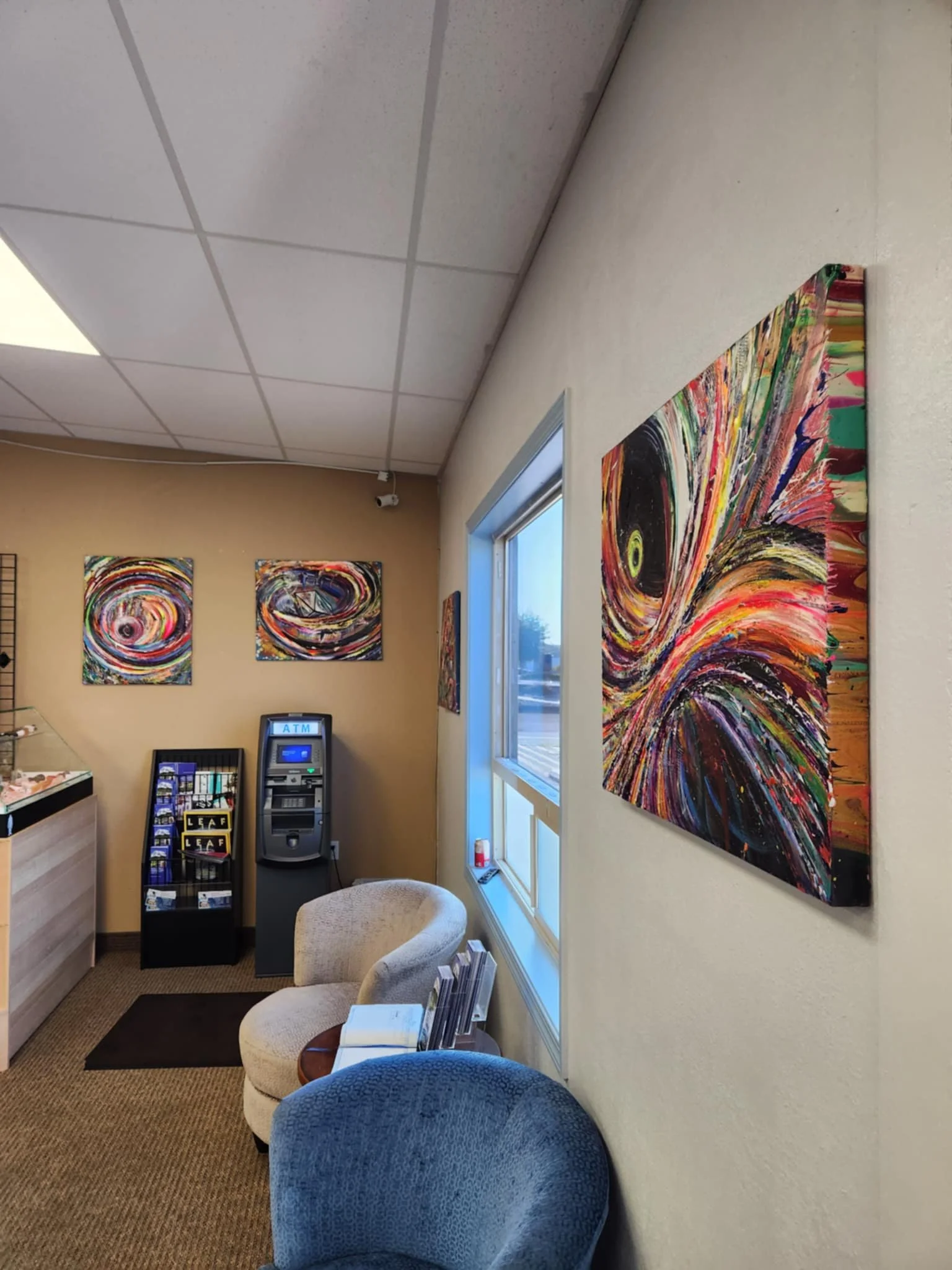 Interior of a waiting area with abstract art on the walls, an ATM, two chairs, and a snack rack. The floor is carpeted, and there's a window on the side wall.