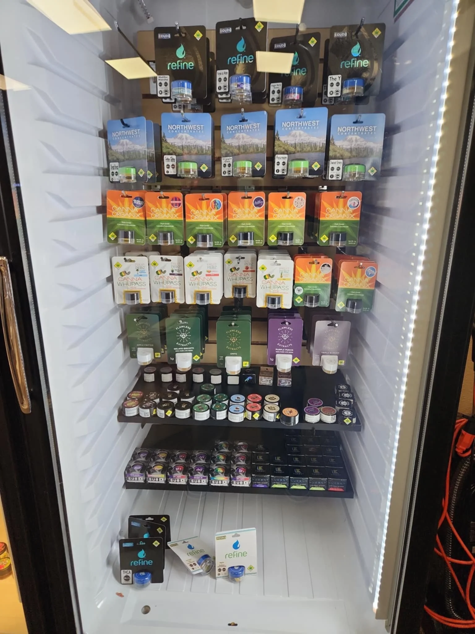 A display refrigerator containing various cannabis products such as vape cartridges and concentrates. The shelves showcase brands like Refine, Northwest, Canna Organix, and Whipass with colorful packaging and labels indicating different strains and t