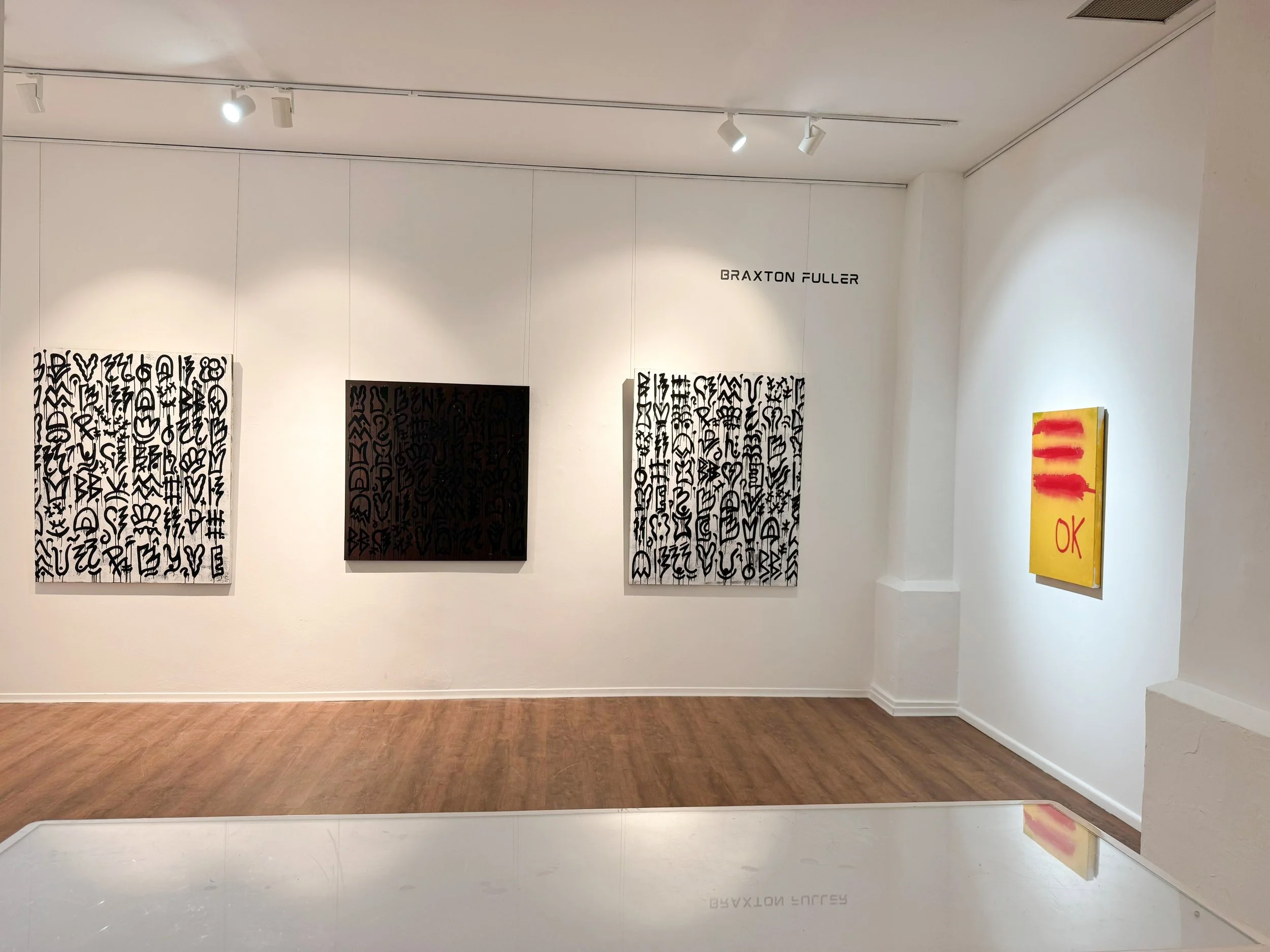 Art gallery wall displaying black and white abstract paintings and a colorful abstract painting, with white wall and wooden floor.