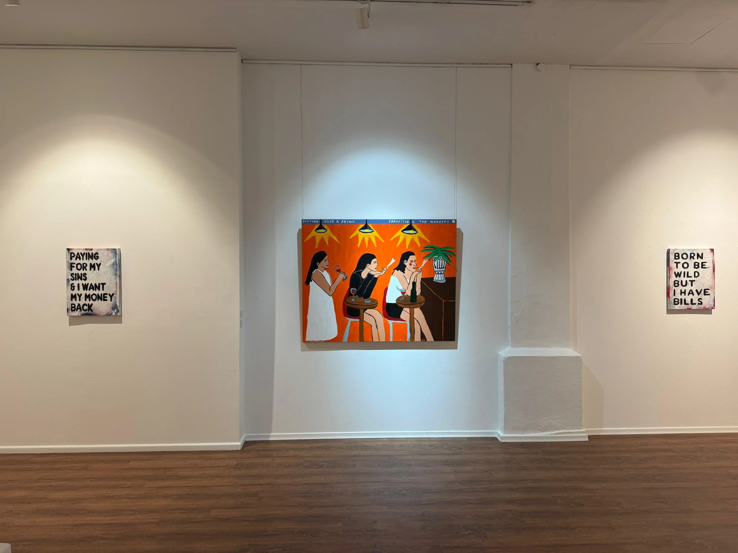 An art gallery wall displays three pieces of artwork: a painting of three women sitting at a bar drinking wine, and two smaller signs with text, one on each side of the painting.