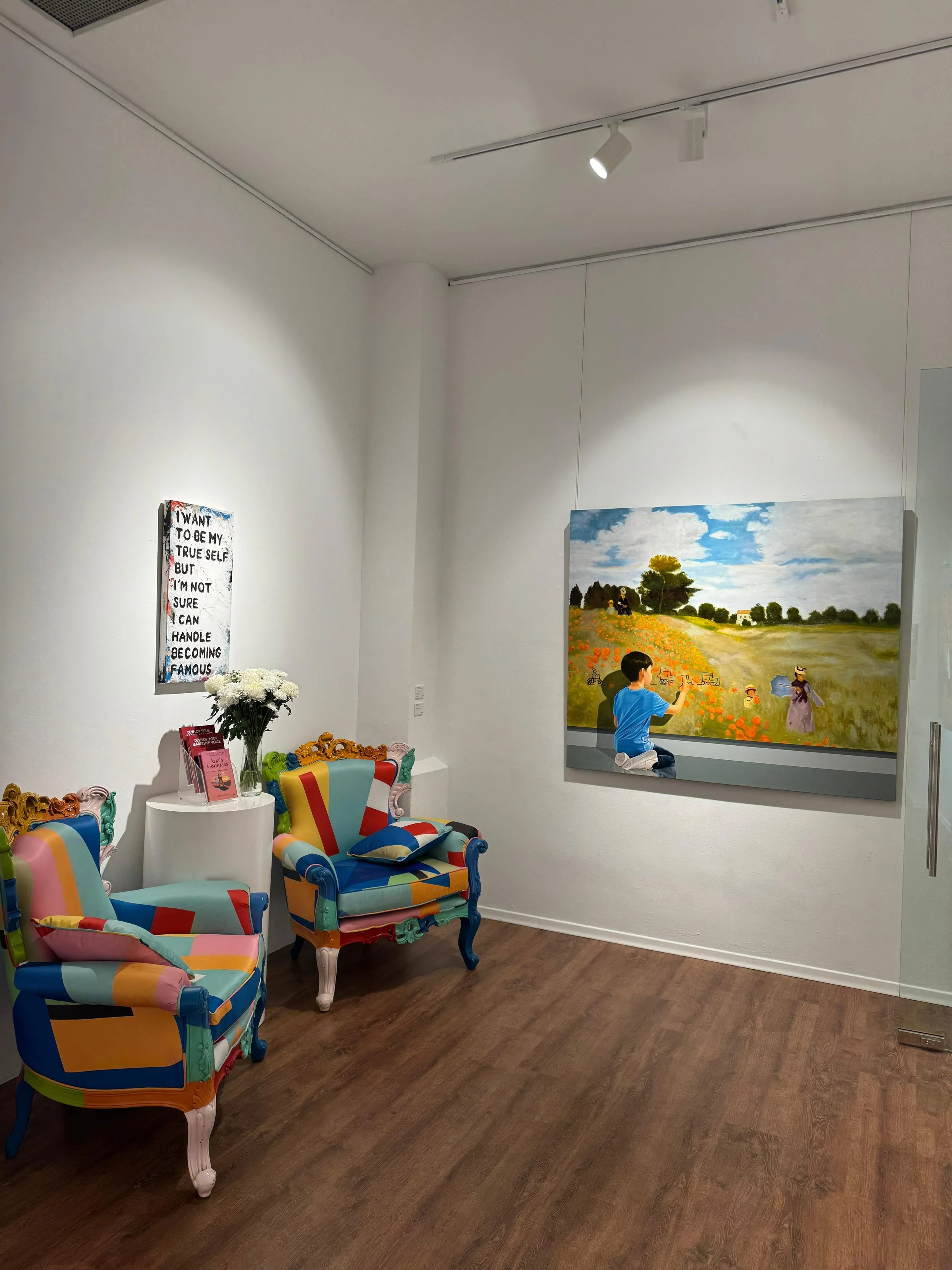 An art gallery with a colorful armchair, a small white table with flowers, all set against white walls. There is a large landscape painting of a field with trees and a blue sky. A small boy is kneeling in front of the painting, engaging with it.