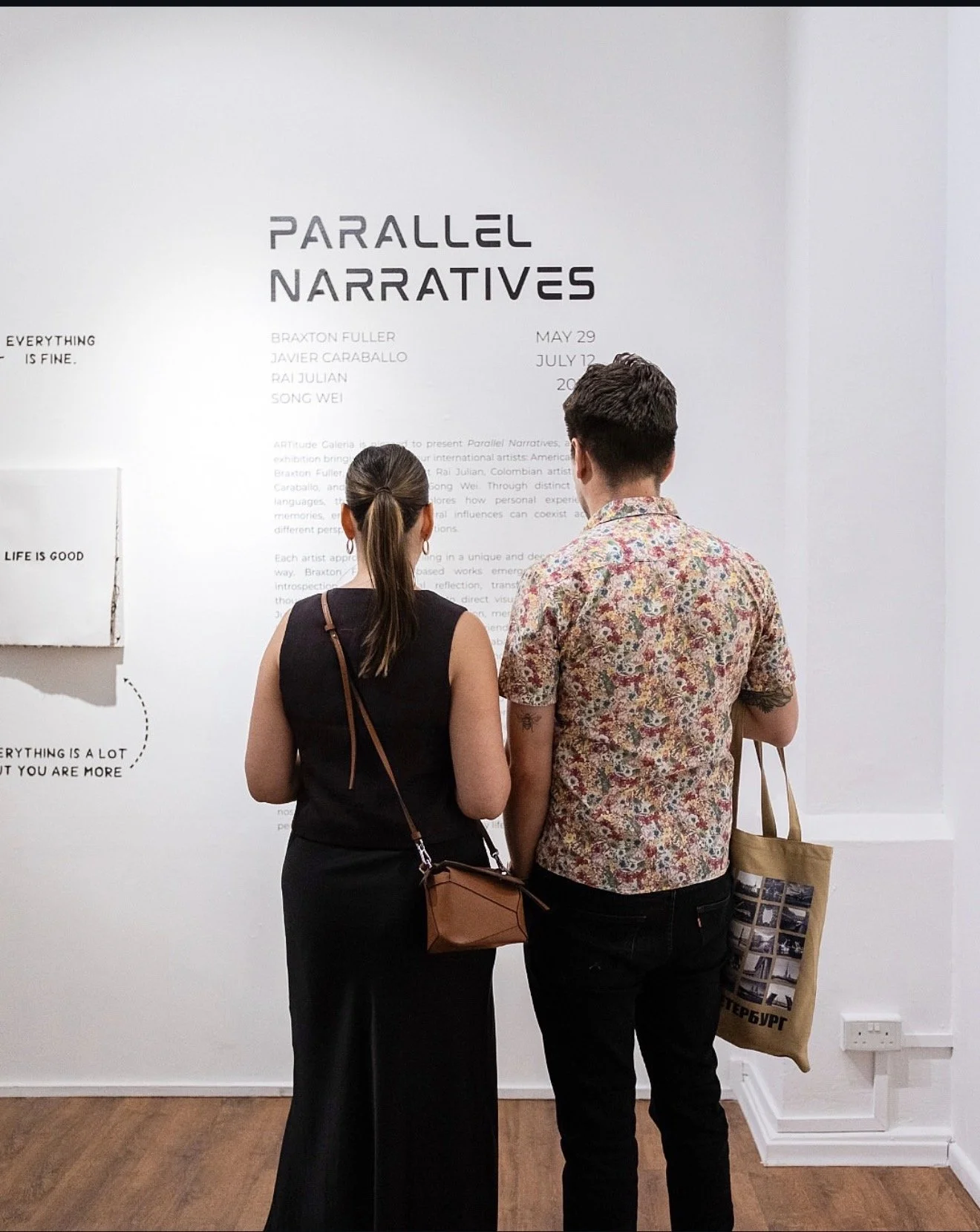 Two people are standing in front of an exhibition wall, reading information. The wall displays the title 'Parallel Narratives' and name and date details of several artists. The woman wears a black dress and has a brown purse, while the man wears a sh