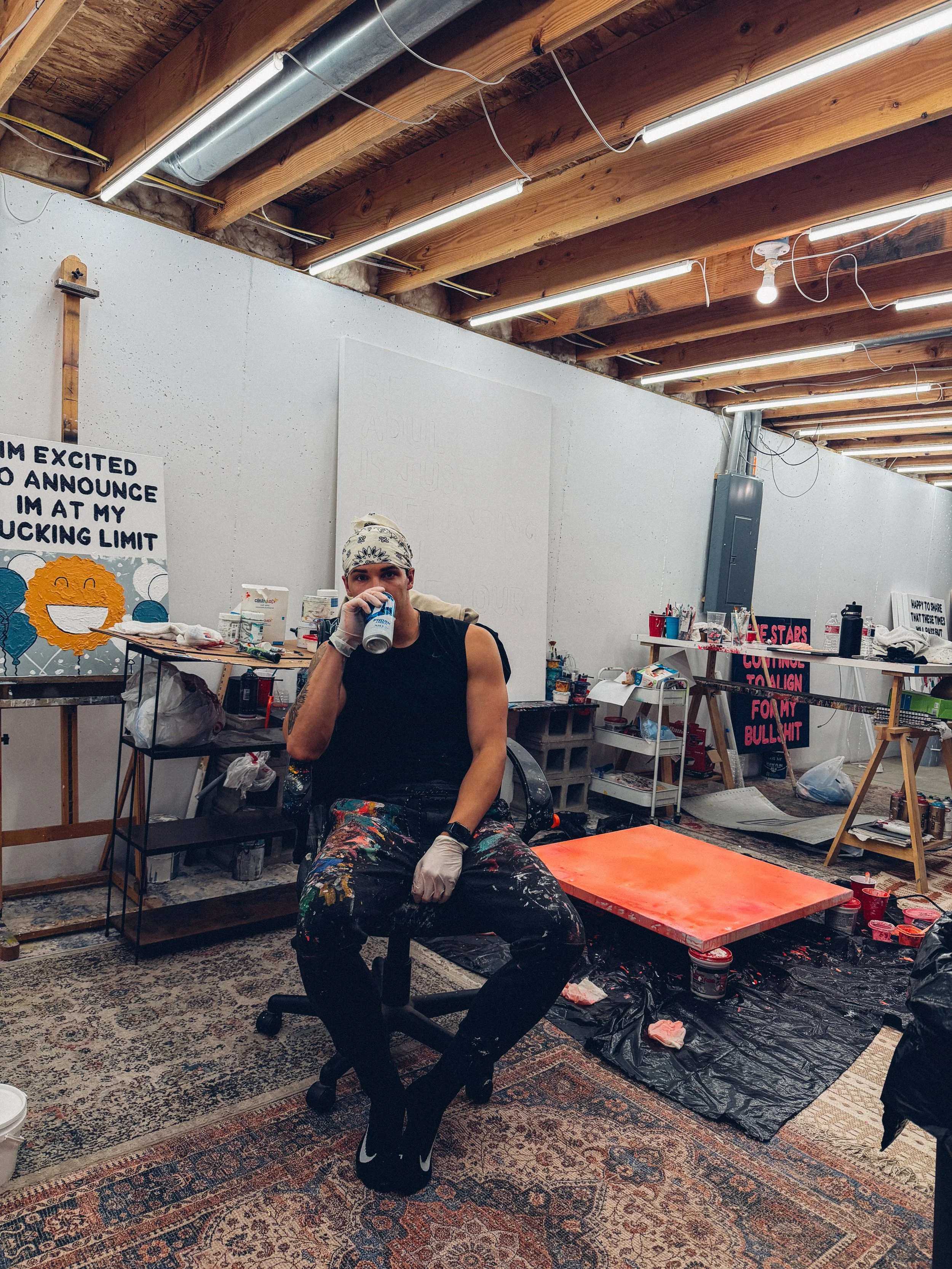 A person with a bandana, black sleeveless shirt, and colorful paint-splattered pants sitting on a black chair, drinking from a can, in an art studio with painting supplies, artwork, and a patterned rug.