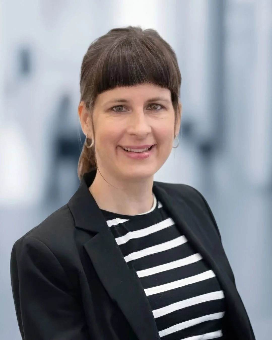 Porträt einer lächelnden Frau mit kurzen braunen Haaren, schwarzen Jacke und gestreiftem Top, im Hintergrund ein moderner, heller Raum.