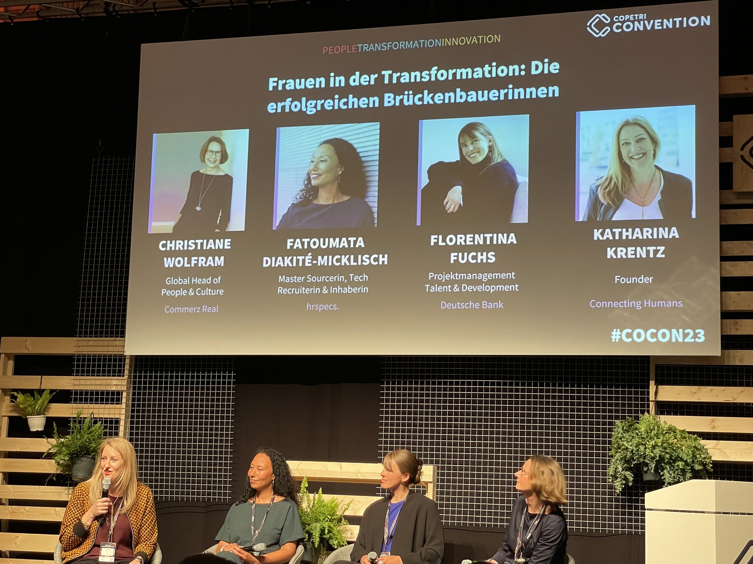 Panel discussion at a conference with four women. The topic of the discussion is &quot;women in transformation: the successful bridge builders&quot;. Photos and names of the participants are shown on the screen: Christiane Wolfram, Fatoumata Diak