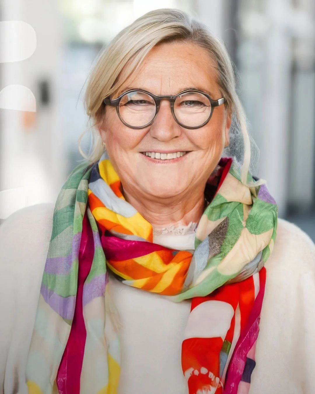 Eine lachende ältere Frau mit grauem Haar, schwarzen Brillen und buntem Schal, sitzend in einem hellen Raum.