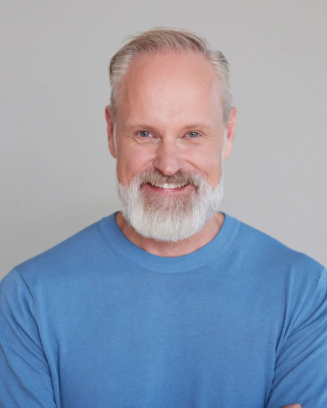 Ein älterer Mann mit grauem Bart und blauen Augen, trägt ein hellblaues T-Shirt, lächelt freundlich, vor einem neutralen Hintergrund.