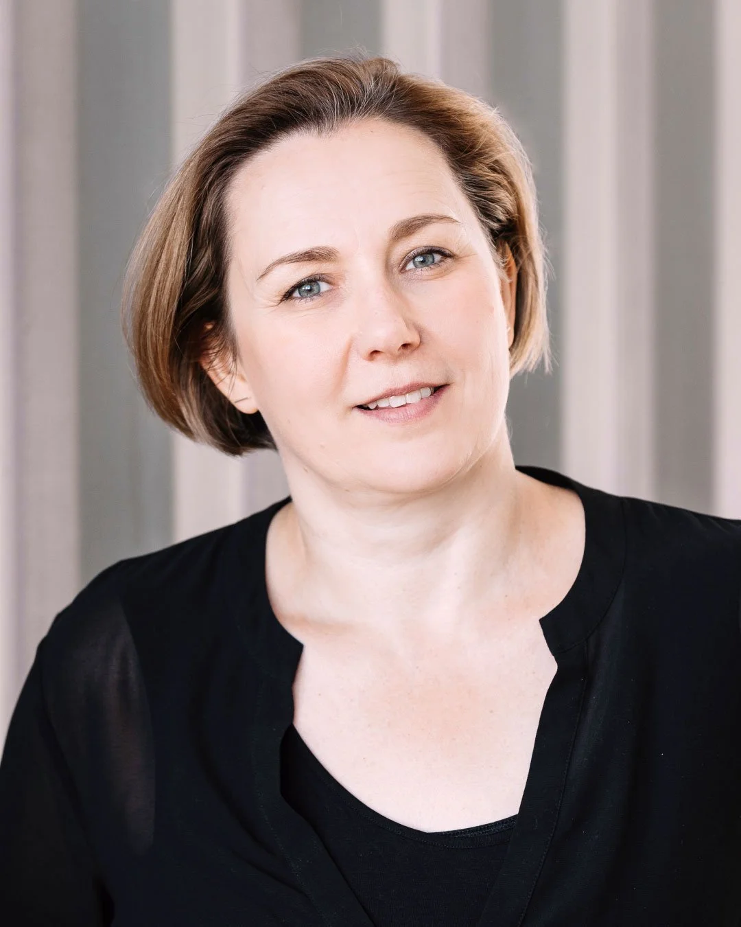 Nahaufnahme einer Frau mit kurzen braunen Haaren, blauen Augen, hellhäutiger Haut, trägt eine schwarze Bluse, im Hintergrund vertikal striped Vorhänge.