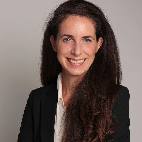Frau im Business-Outfit mit langem braunem Haar, die freundlich in die Kamera lächelt. Sie trägt ein weißes Hemd und einen schwarzen Blazer. Neutrale, graue Hintergrundfarbe.
