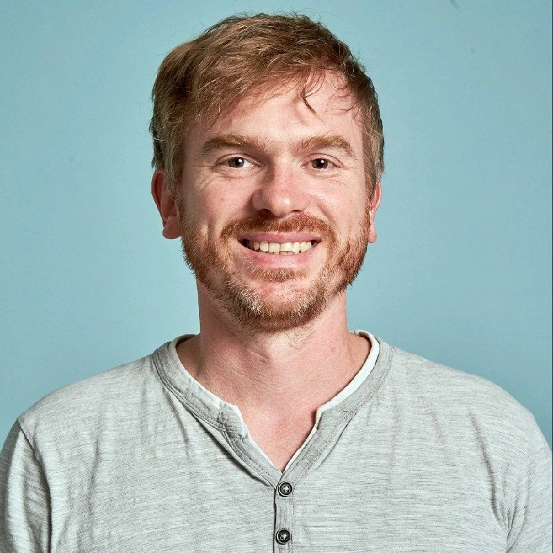 Ein lächelnder Mann mit kurzen Haaren und Bart in einem grauen Hemd vor einem hellblauen Hintergrund.