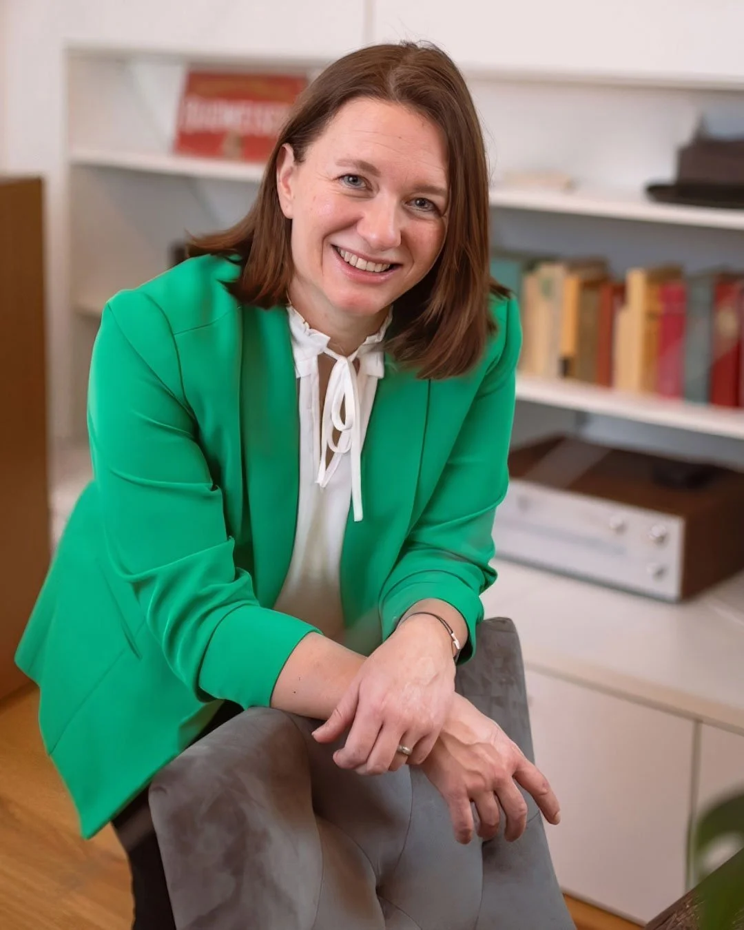 eine lächelnde Frau mit braunen Haaren, grüner Jacke und weißem Hemd sitzt an einem Tisch in einem Büro.