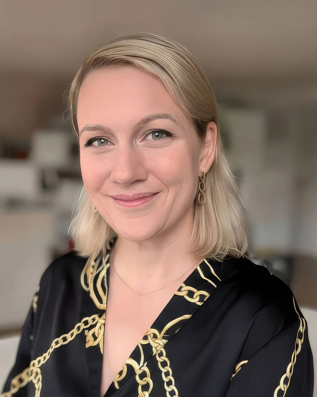 Porträt einer Frau mit blondem, schulterlangem Haar, schwarzen Top mit goldener Kettchenmuster, freundlichem Lächeln, Eheringen und Ohrringen vor einem unscharfen Innenraum