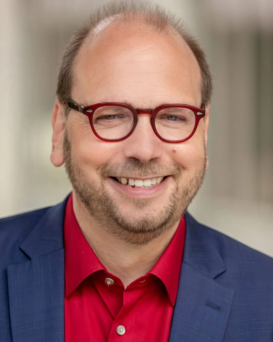 Nahaufnahme eines Mannes mit Brille, bärtig, im Anzug, lächelnd.