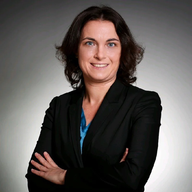 Frau in einem schwarzen Blazer mit den Armen verschränkt, lächelnd, mit dunklem, schulterlangem Haar vor einem grauen Hintergrund.
