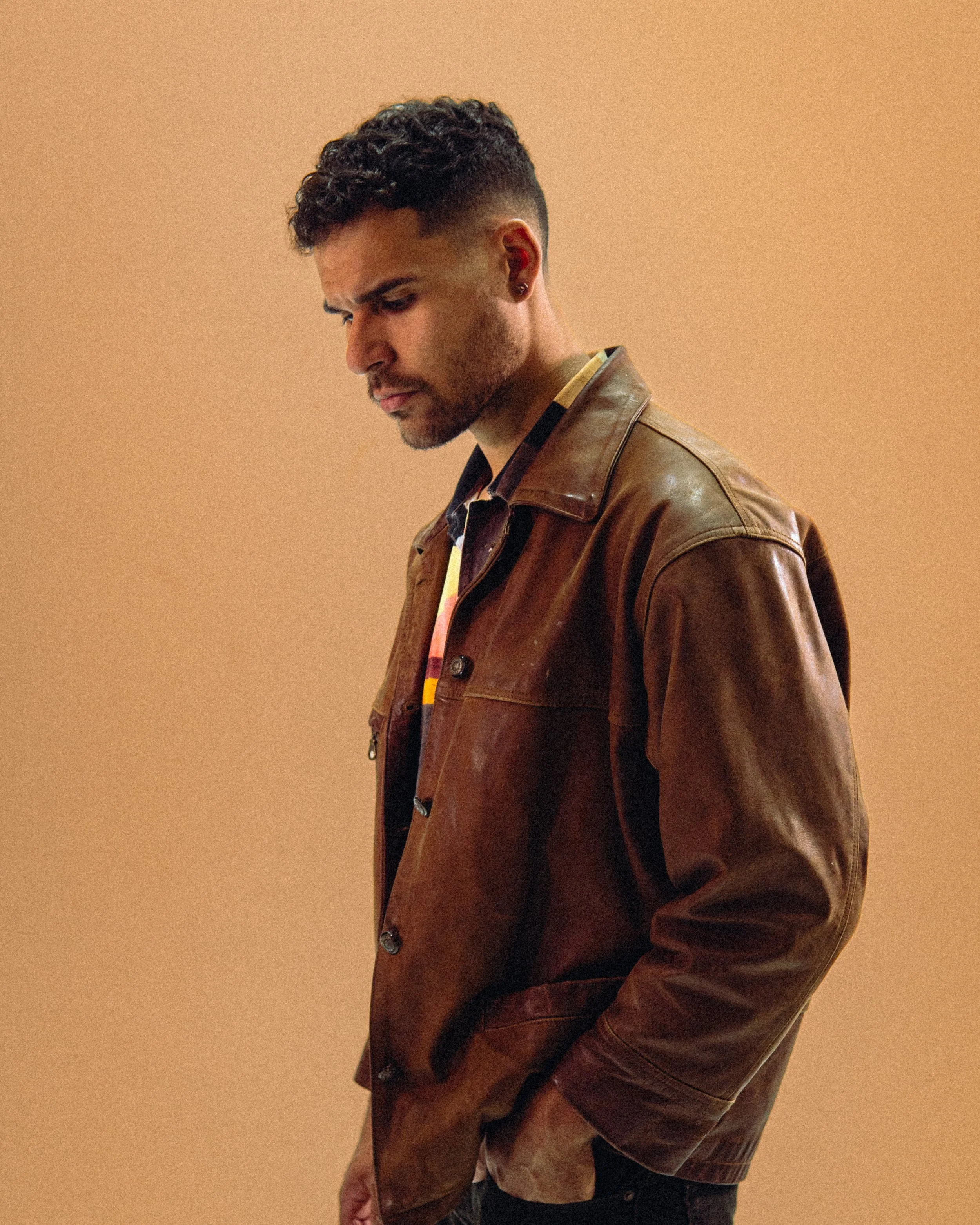 Reece Forward, a man with curly hair, a beard, and earrings looking down, wearing a brown leather jacket, against a plain beige background.