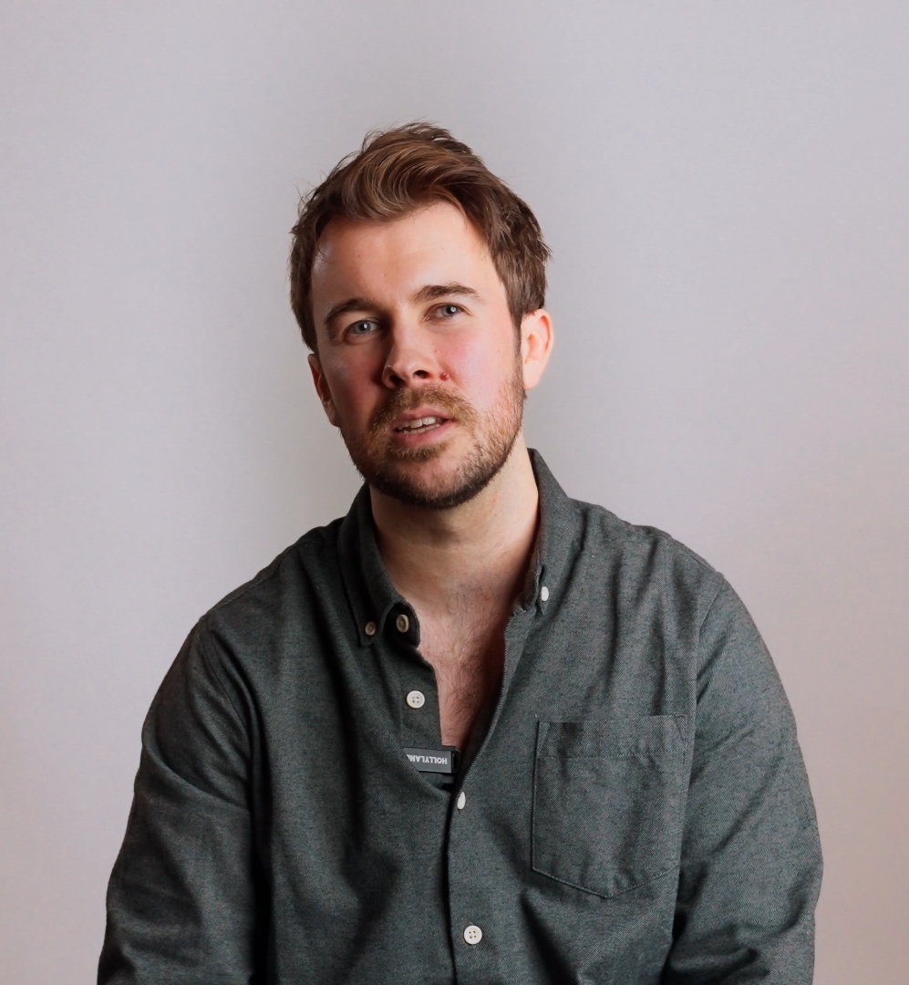 A man with short brown hair and a beard, wearing a dark gray button-up shirt, sitting against a light gray wall.