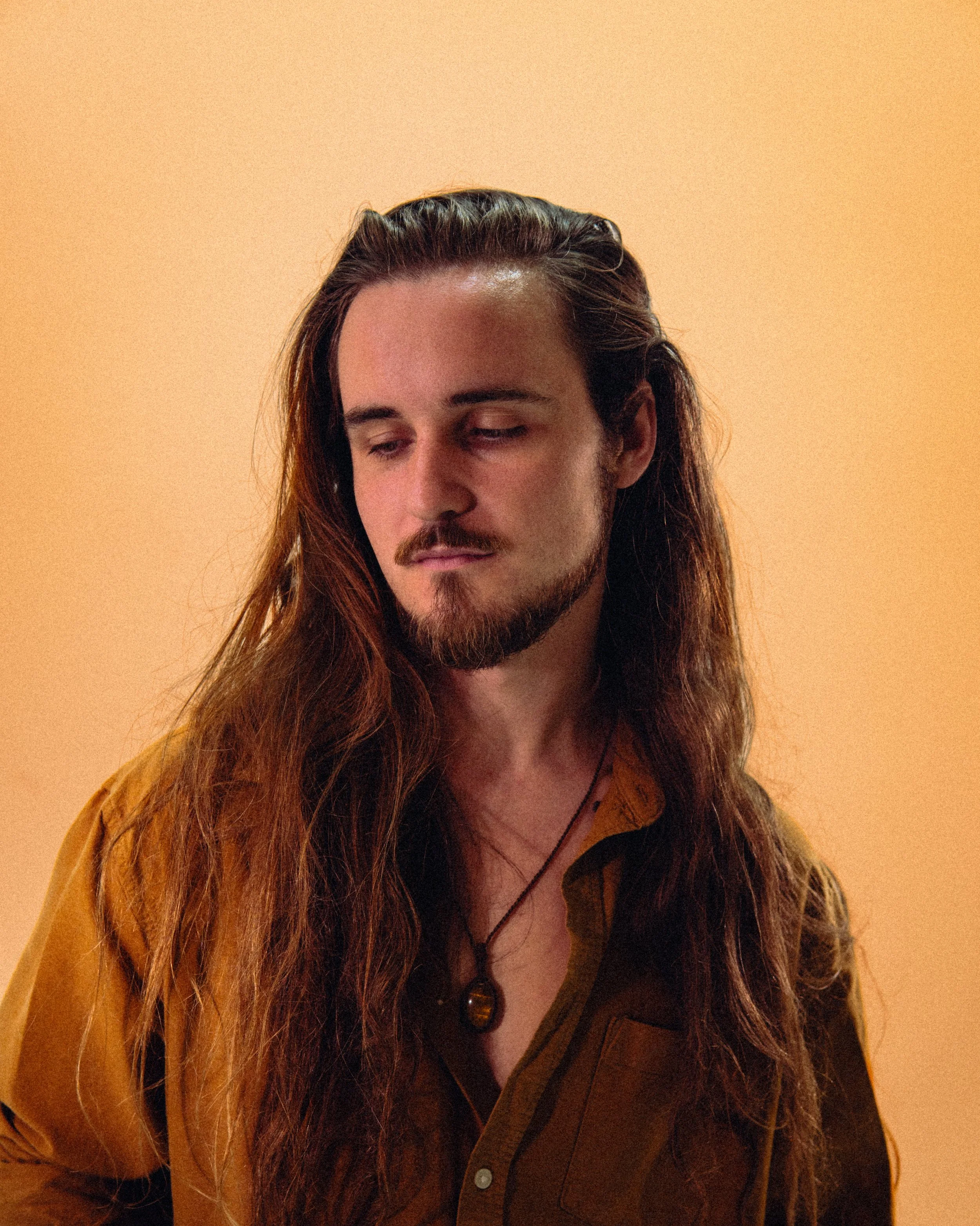 A man with long brown hair, a beard, and a mustache looking downward, wearing a mustard-colored shirt and a necklace with a pendant, against a plain orange background.