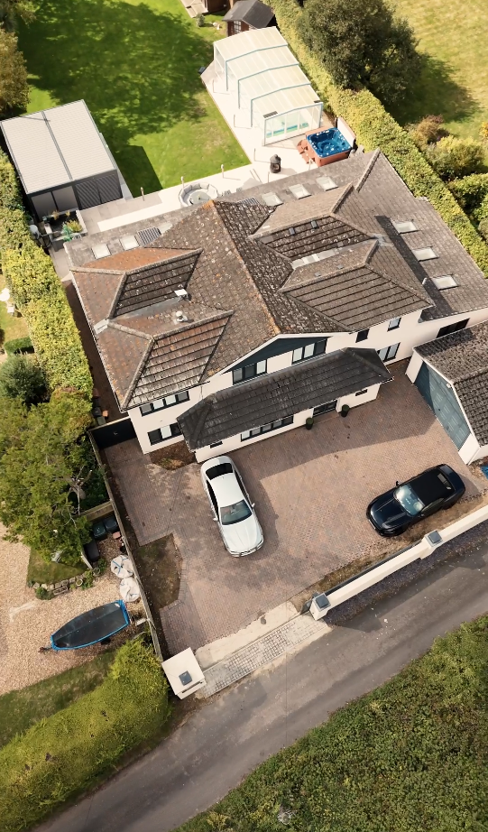 Aerial view of a two-story house with a tiled roof, a driveway with two parked cars, a small boat, a backyard with a shed, a greenhouse, a hot tub, and surrounding greenery.