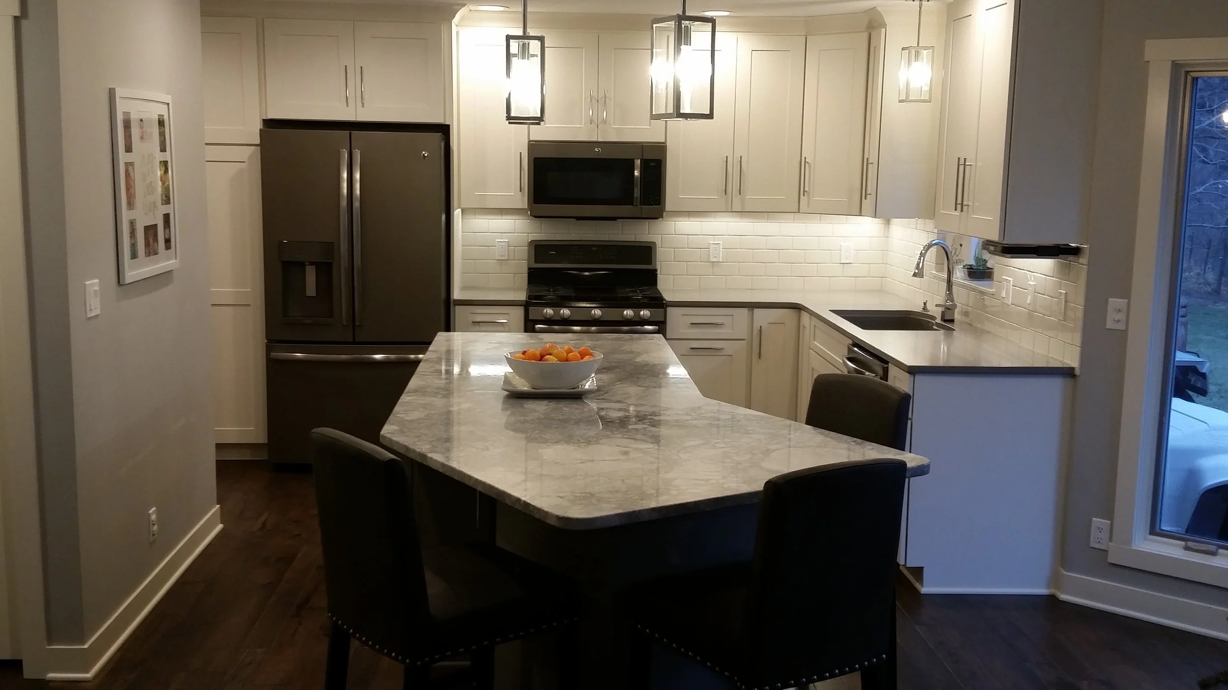 Modern kitchen with white cabinets, stainless steel refrigerator, microwave, and oven, granite countertop island with a bowl of oranges, dark hardwood flooring, pendant lighting, and a sliding glass door to an outdoor area.