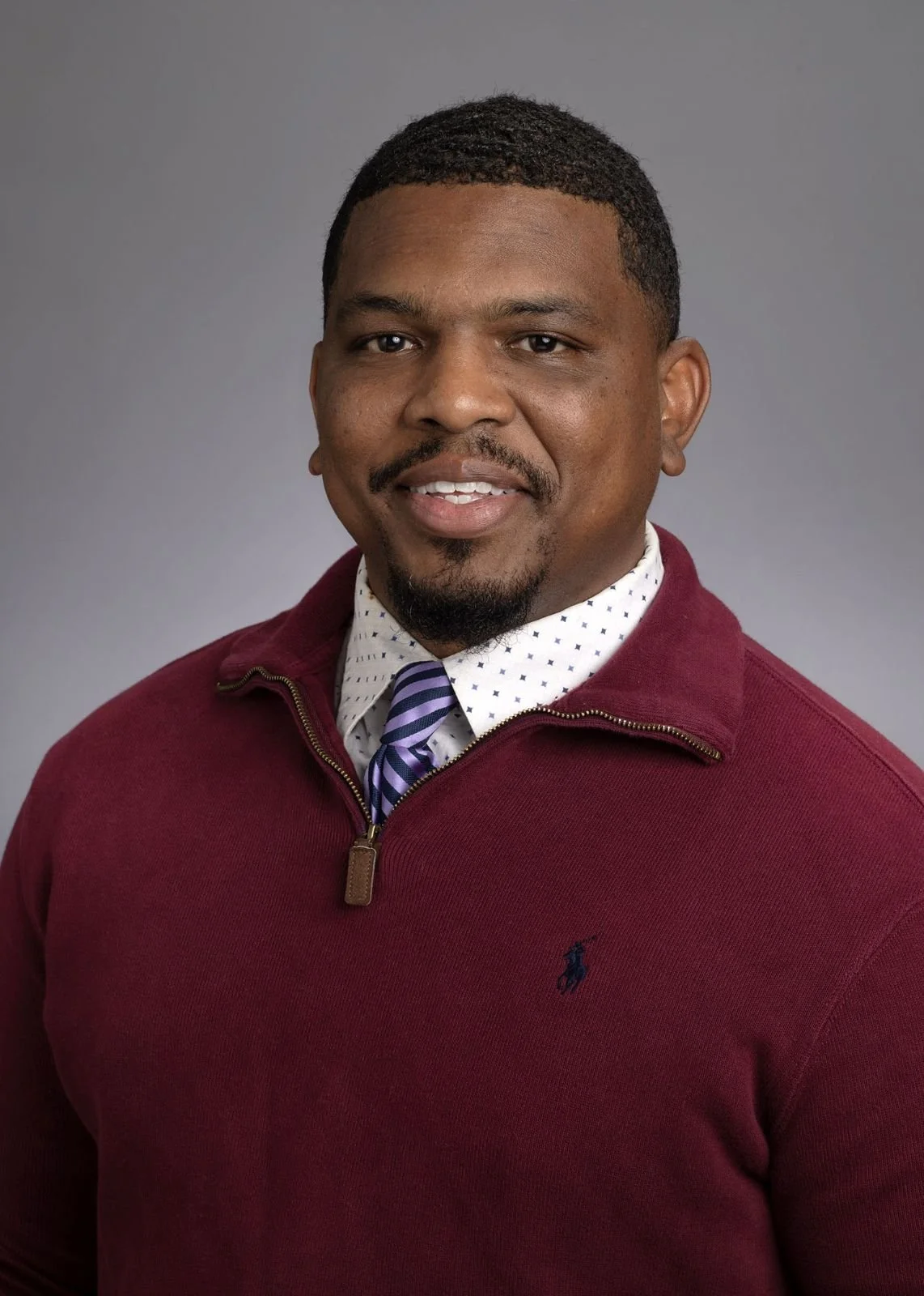 Portrait of a man with short curly black hair, a goatee, wearing a red sweater with a zipper, a white shirt with blue polka dots, and a striped purple and blue tie, against a gray background.