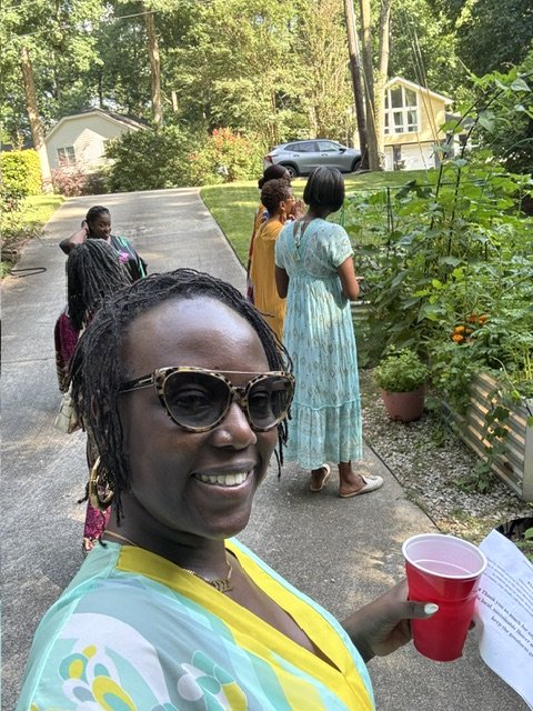 A woman taking a selfie outdoors with a group of women in the background, some looking at plants, in a garden or backyard setting.