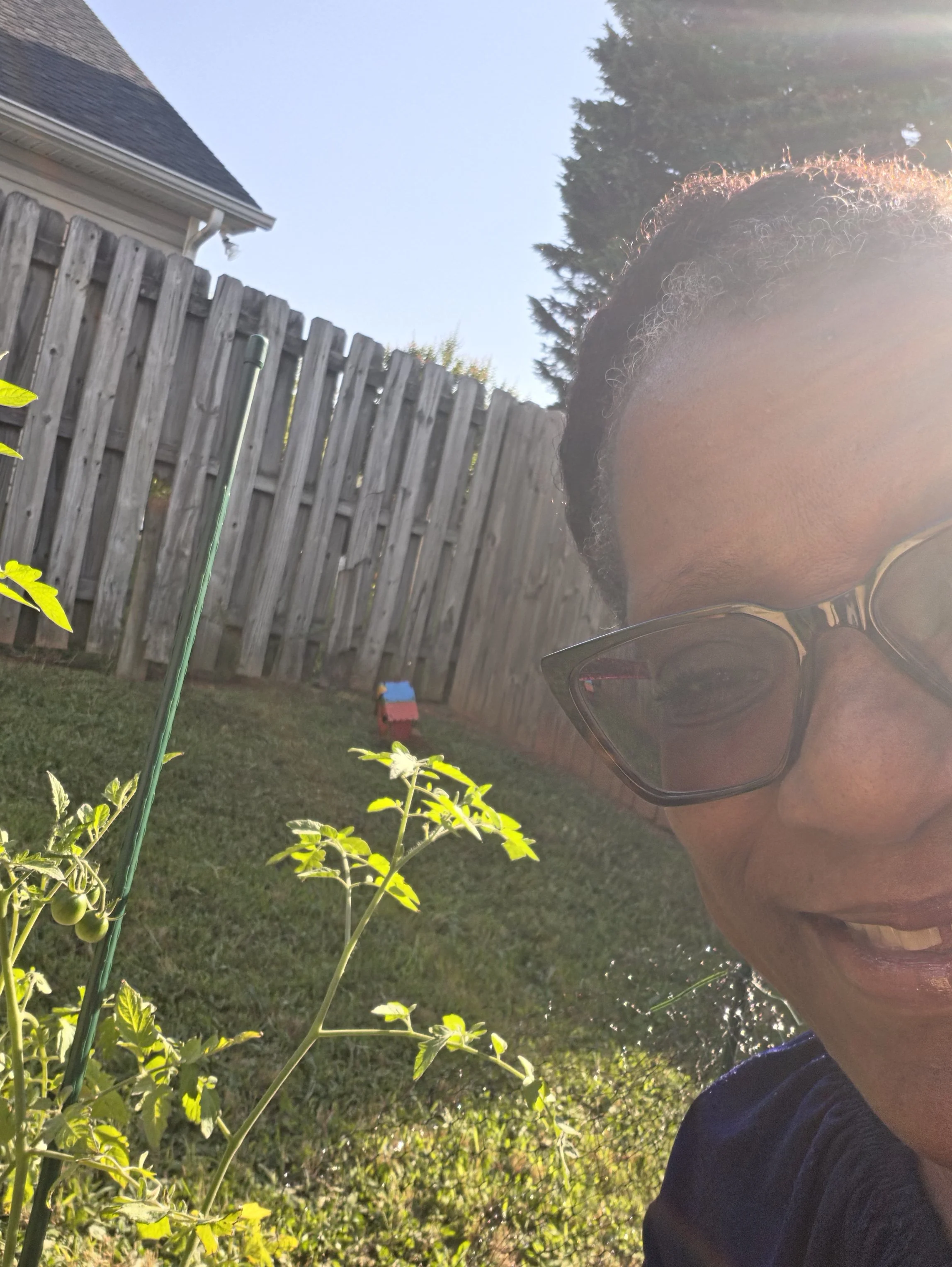 A person with glasses taking a selfie in a backyard with a wooden fence, a house with a gable roof, and a tall tree in the background during daytime. The person is partially cut off on the right side of the image.