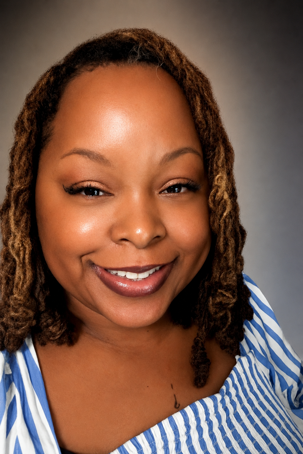 A smiling woman with medium brown skin and shoulder-length, curly hair, wearing a blue and white striped shirt against a gray background.