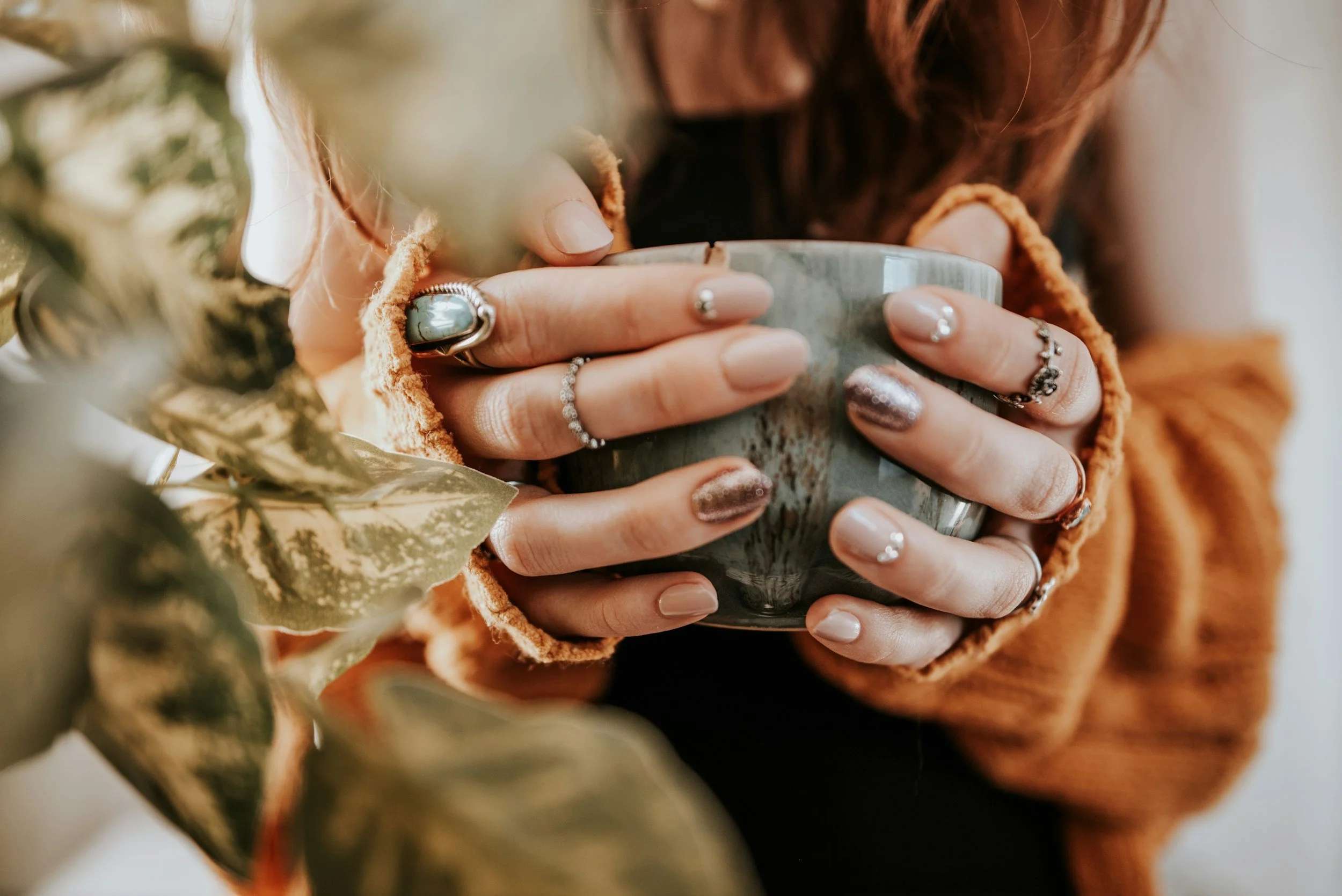 Mains tenant une tasse, ongles vernis, vêtements en tricot orange, plantes floues au premier plan.