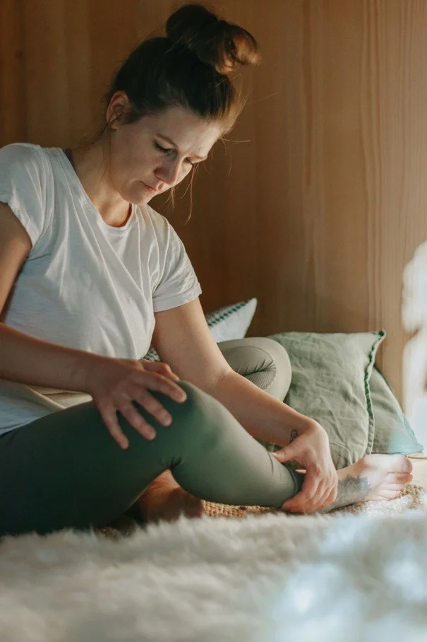 Femme assise sur un lit, regardant ses jambes, portant un t-shirt blanc et un pantalon vert, dans une chambre avec des oreillers verts et une couverture en fourrure blanche.